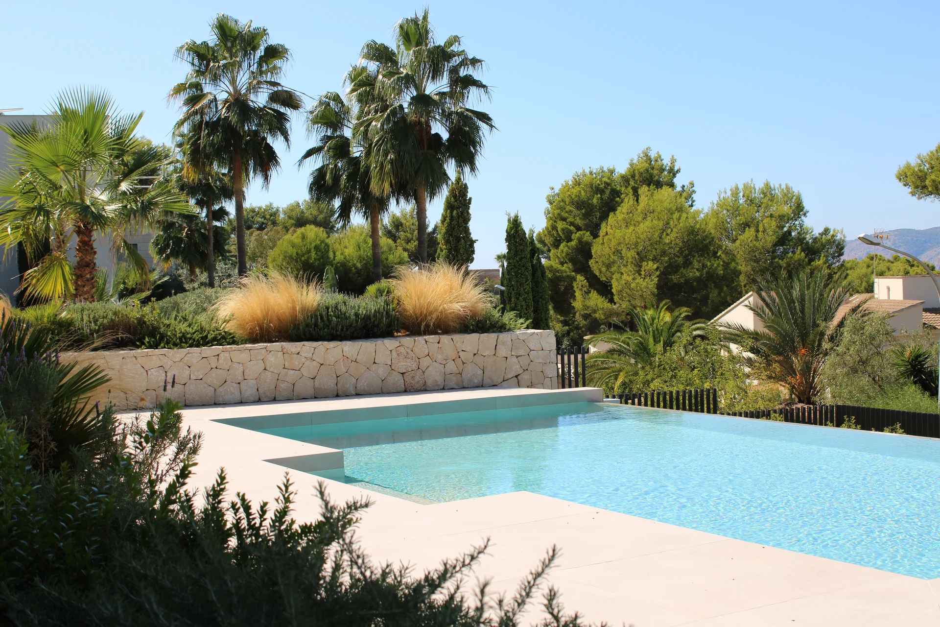 Terraza de piscina con muro tradicional de piedra seca mallorquina, gramíneas ornamentales y palmeras - S'Estepa Design