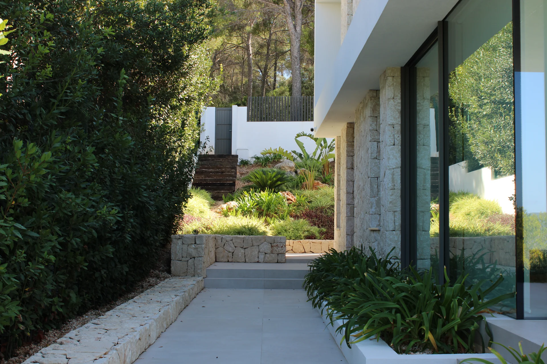 Corredor lateral de piedra con escalones, fachada moderna de mampostería y jardinera hacia el talud - S'Estepa Design